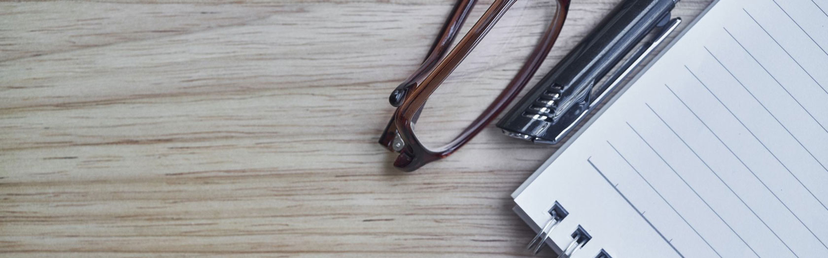 glasses, with pen and pad of paper on wood desk